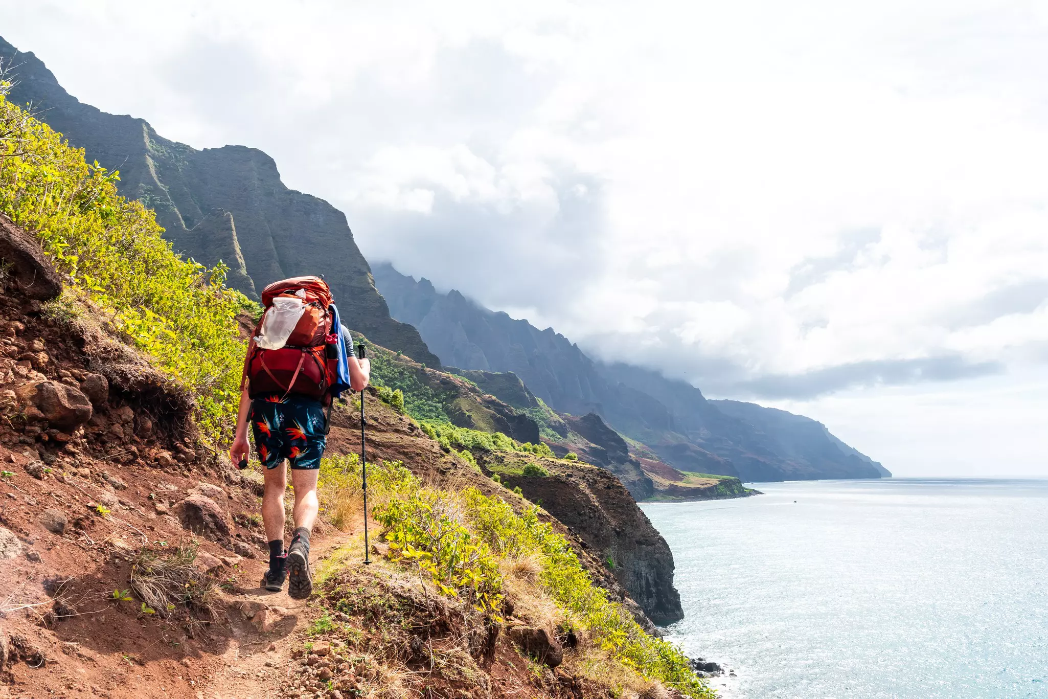 A man walks along a narrow path along a cliff. Mountains rise to his right, and the sea is to his left.