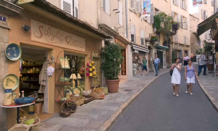 People strolling a sloping old-town street past a pottery shop displaying colorful glazed plates and baskets