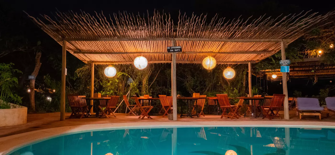 A pool at night lit by lights over the nearby tables and chairs.