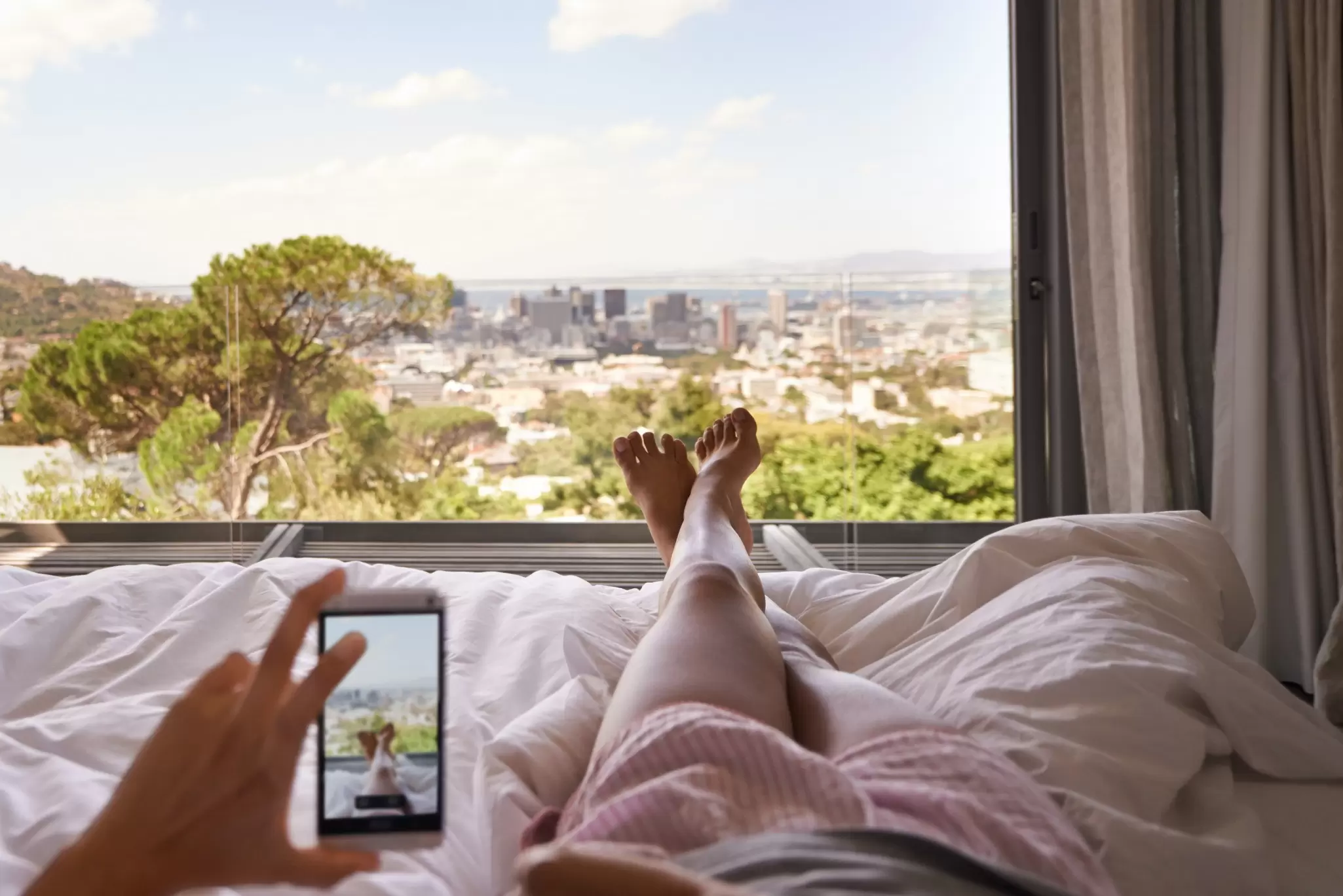 View from a bed of bare feet and a phone photographing a sunny hillside city and harbour through a large window