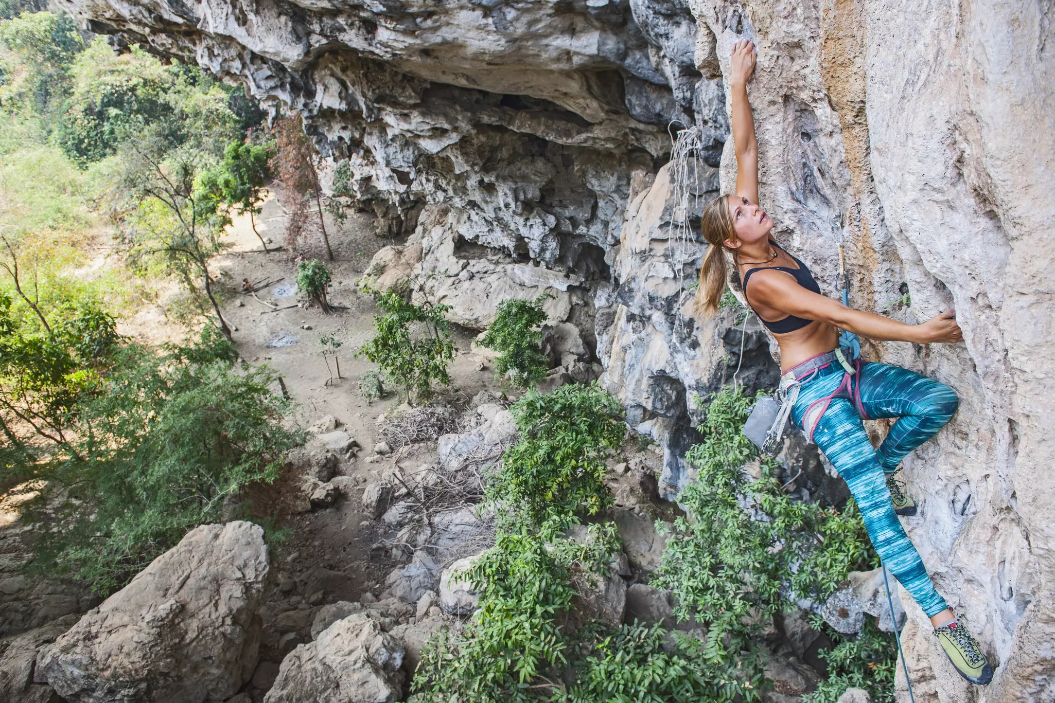 A female climber ascends a rock face in Tha Khaek, Laos, with the rocky ground far below her.