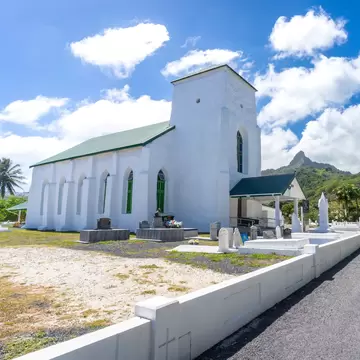 White-washed coral church with green roof and graves, a road beside it and jagged peaks behind, Rarotonga