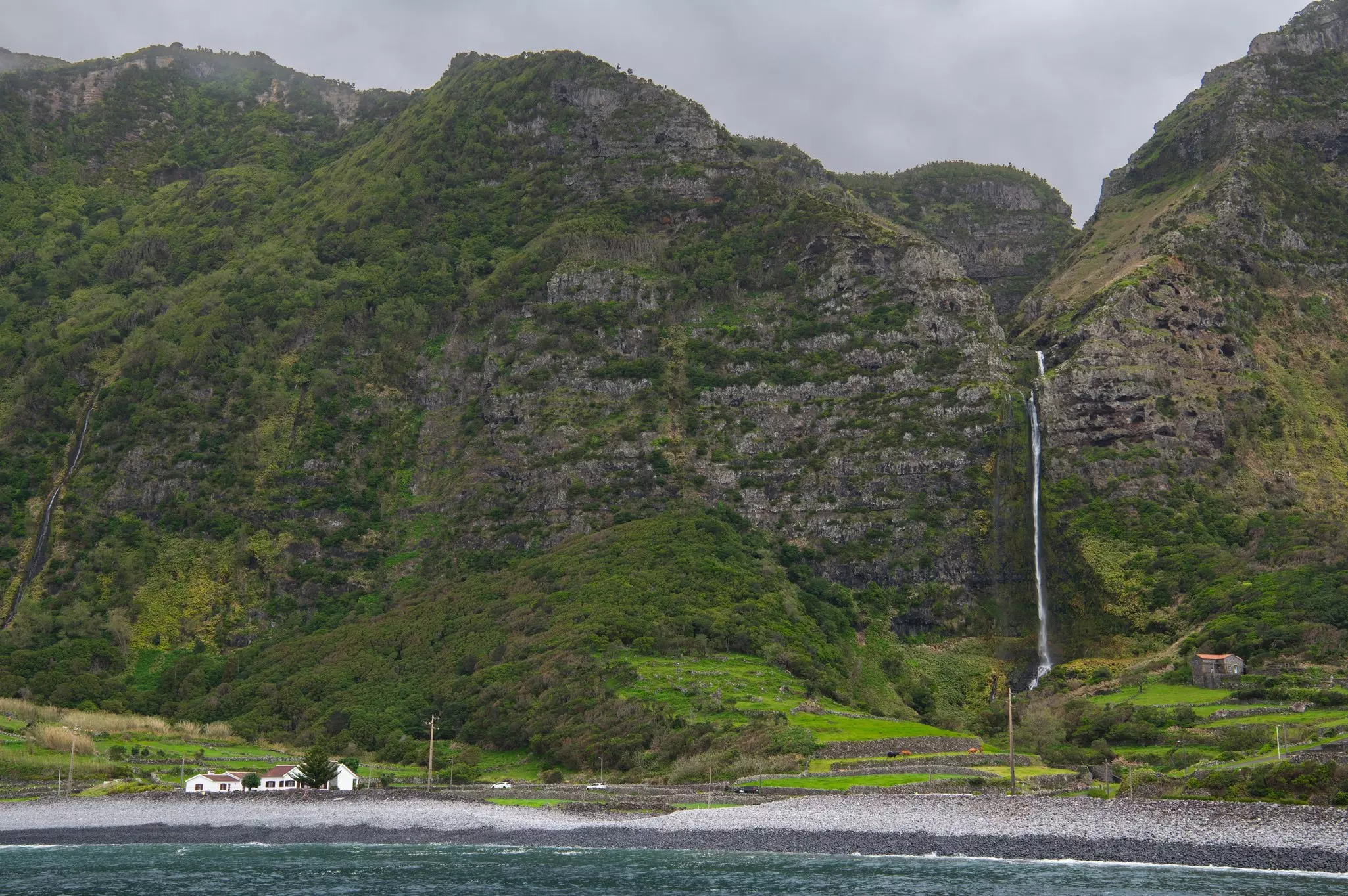 A small waterfall falls down lush cliffs in front of a black-sand beach on the coast. A small white house is visible at the lower left of the image.