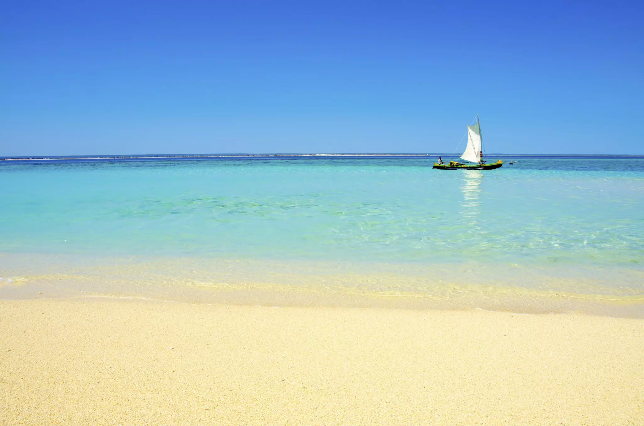 A small wooden sailboat in turquoise ocean just off the shore of a sandy beach