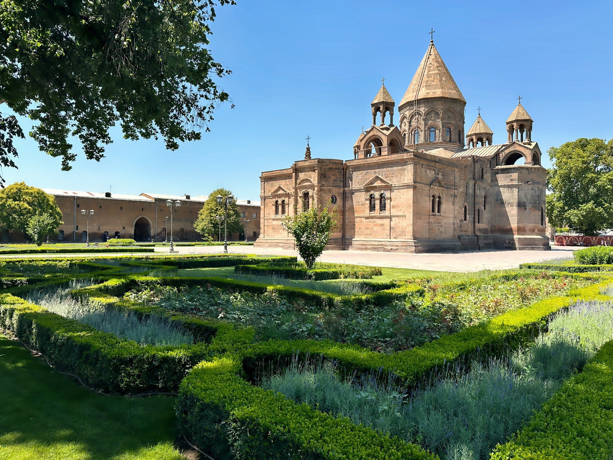 A domed stone cathedral with a conical roof behind manicured hedge gardens.