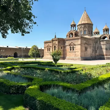 A domed stone cathedral with a conical roof behind manicured hedge gardens.