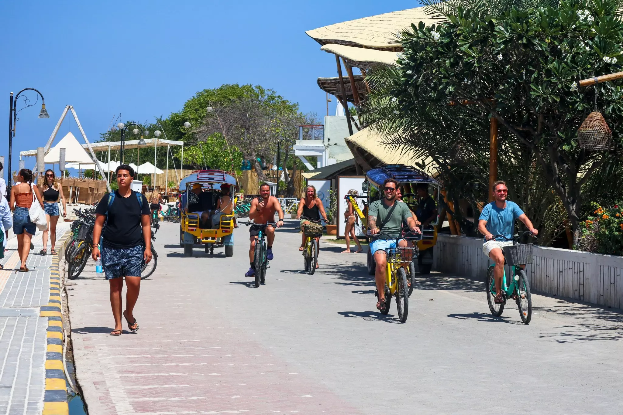 Tourists cycling and walking past a restaurant and shops on a sunny day with clear blue skies