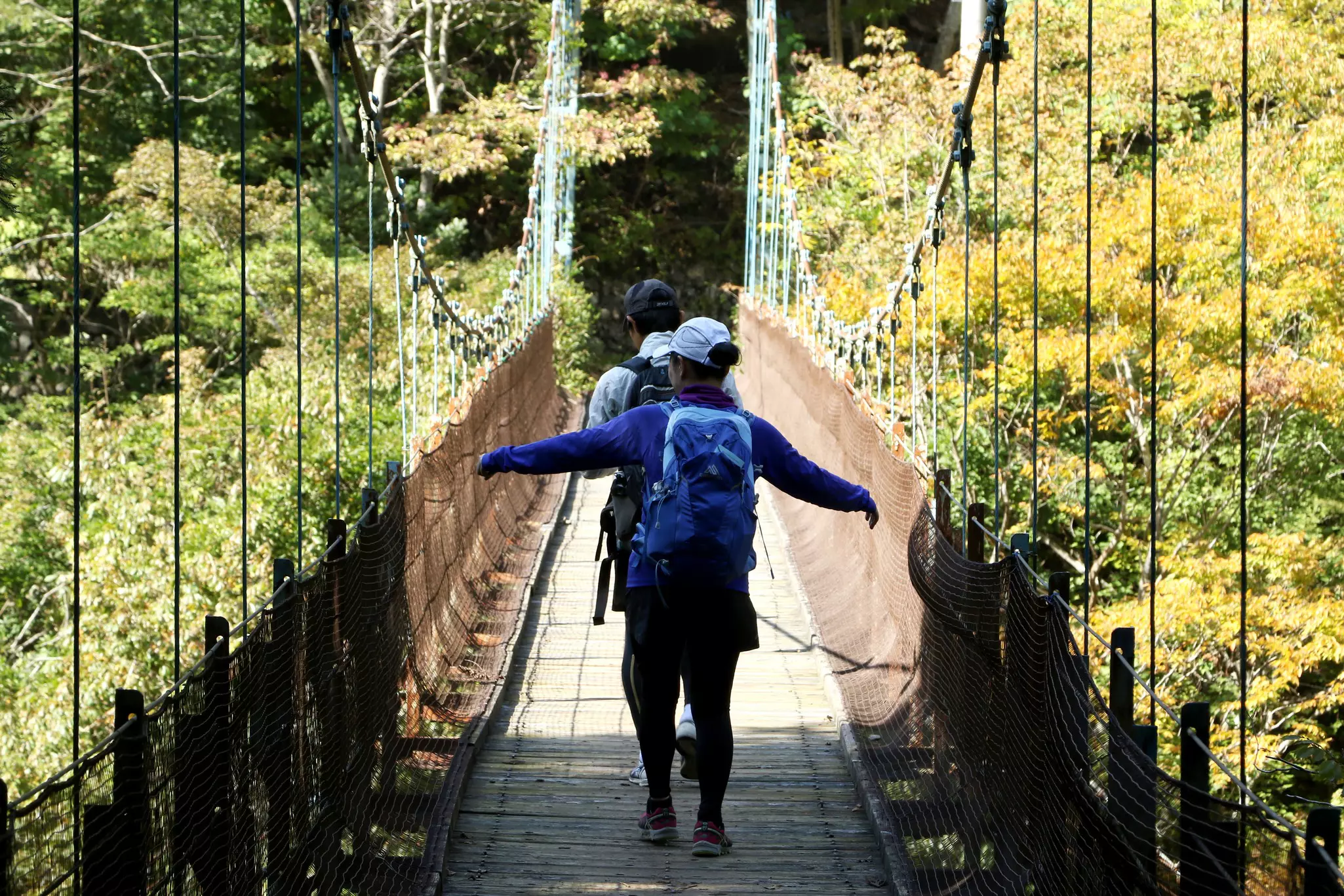 Two people with backpacks and wearing ballcaps walk single file on a suspension bridge on a sunny day; the person walking in back has their arms out to the side.
