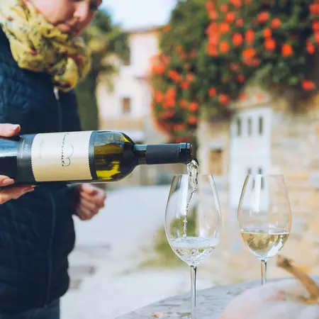 A person pours white wine from a bottle into a clear glass; another glass with white wine is to the side.