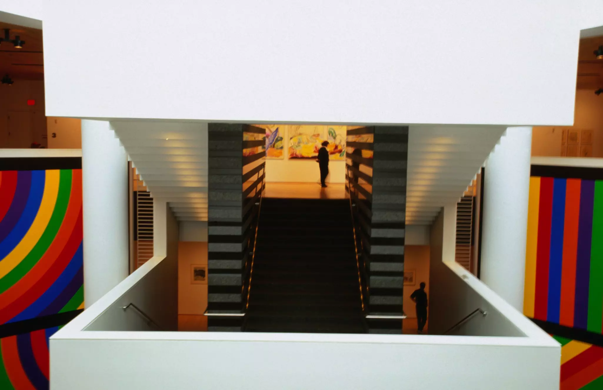 A person in silhouette in an entryway to a gallery at the end of a white staircase; colorful artwork is on either side.