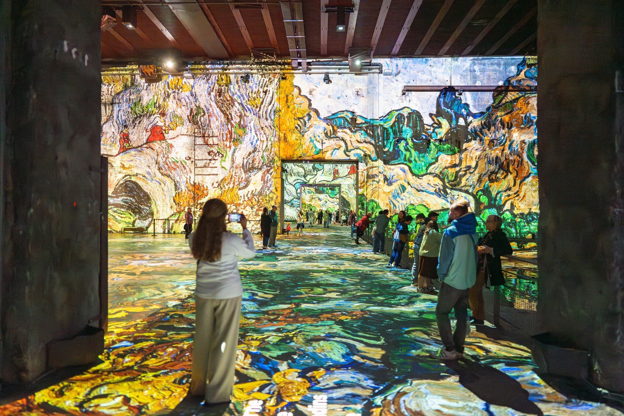 Visitors admiring large-scale digital projections of Van Gogh artworks at Les Bassins des Lumières, featuring vibrant colors and reflective floors in Bordeaux, France.