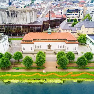 White building with an orange tiled roof and green cupola behind rows of trees and a flower-edged lakeside path.