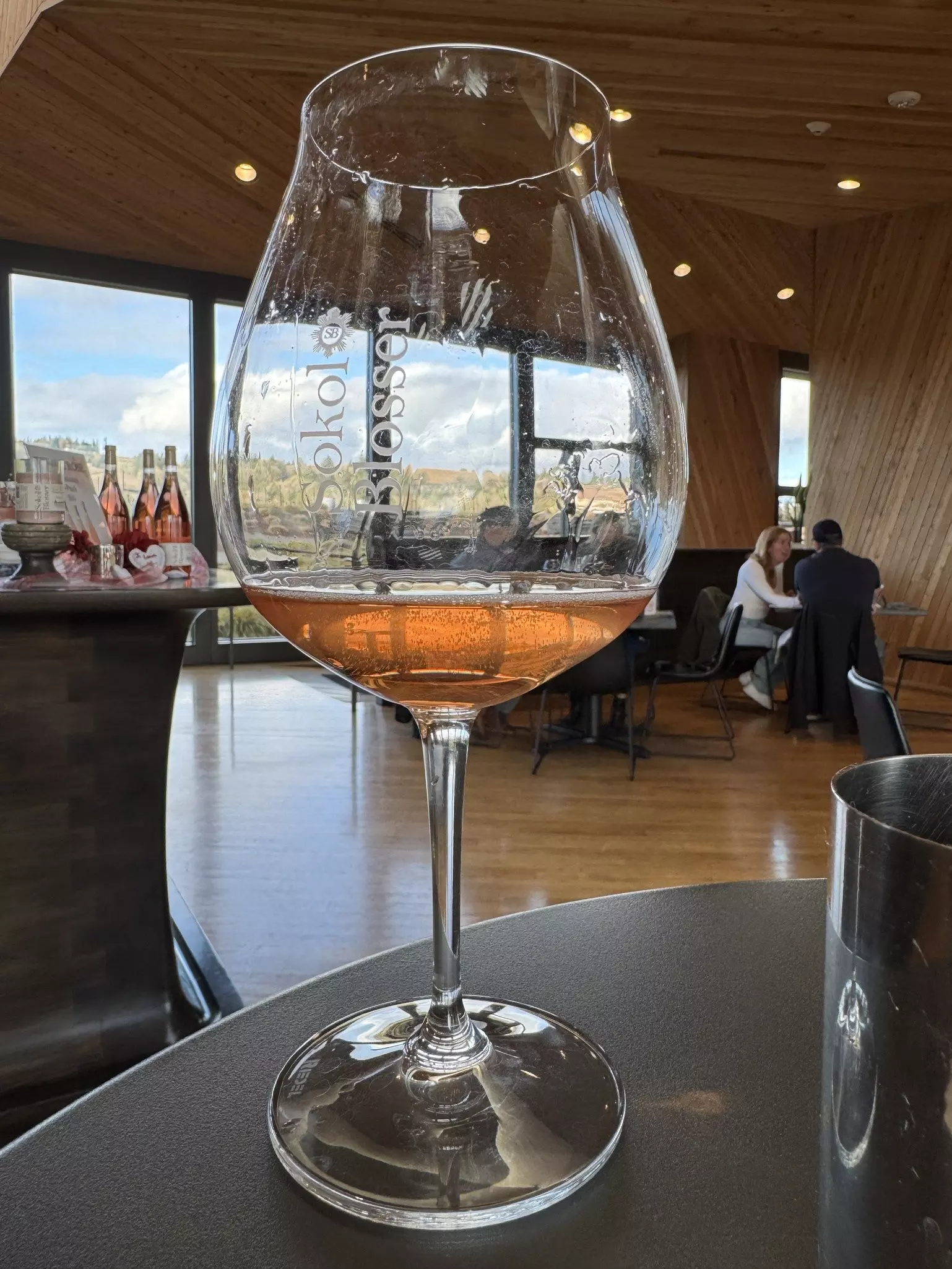 A wine glass with a small amount of pink liquid on an indoor table. People can be seen sitting at tables in the background, and trees and clouds can be seen through large windows on a sunny day.