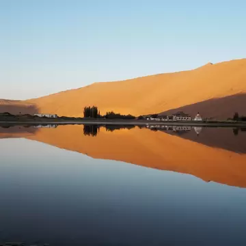 Tall orange sand dune in low evening sun, mirrored in a calm lake with a cluster of white buildings and trees on the shore.