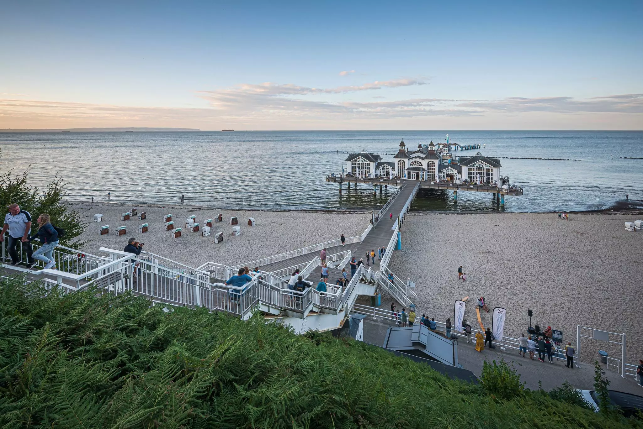 A boardwalk over a beach leads to a white and gray building with a pier stretching into the sea beyond.