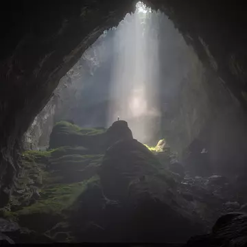 A person stands in a shaft of light flooding downwards into a vast cave system.