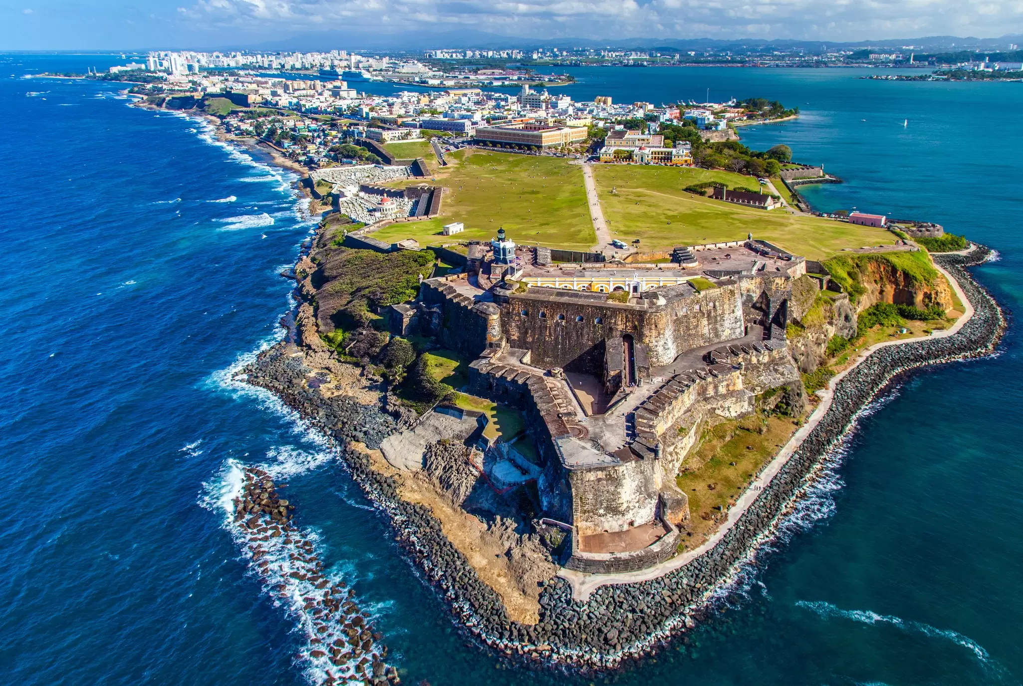 An aerial view of a historic fort at the tip of a peninsula. A large city can be seen in the distance, and white-capped waves break along the shore.