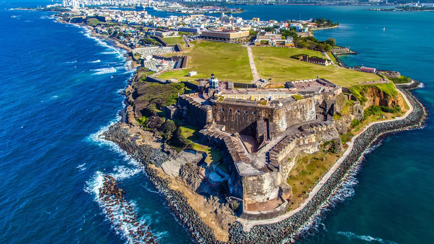 An aerial view of a historic fort at the tip of a peninsula. A large city can be seen in the distance, and white-capped waves break along the shore.