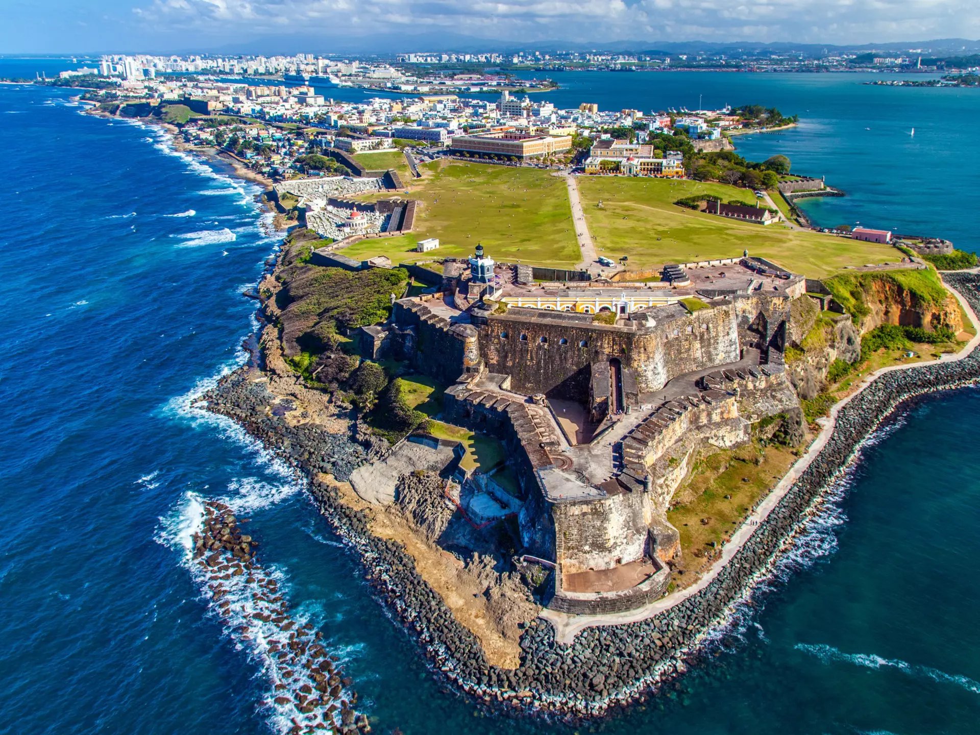An aerial view of a historic fort at the tip of a peninsula. A large city can be seen in the distance, and white-capped waves break along the shore.