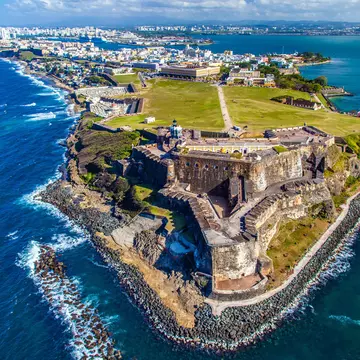 An aerial view of a historic fort at the tip of a peninsula. A large city can be seen in the distance, and white-capped waves break along the shore.