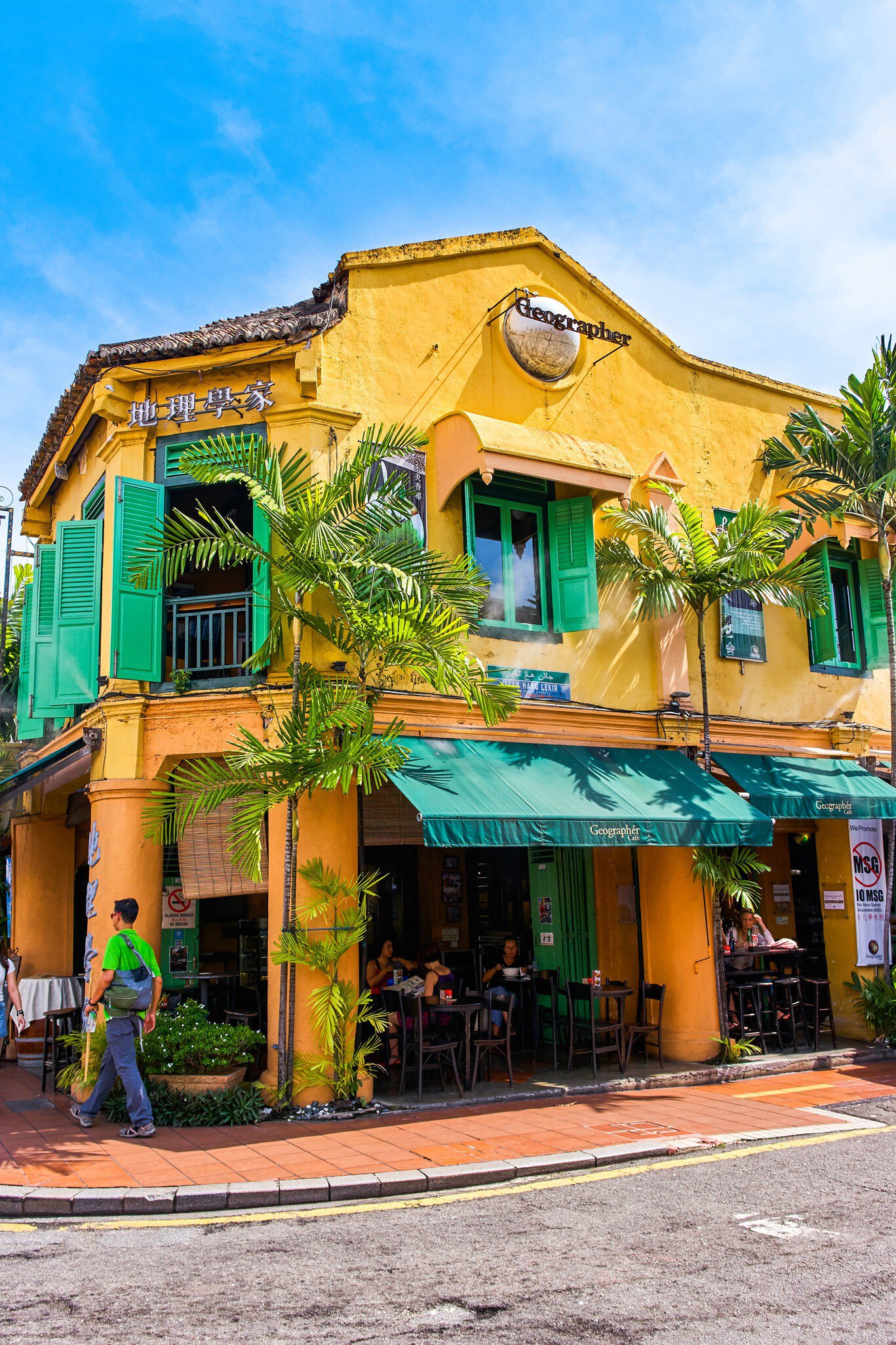 Yellow two-storey shophouse with green shutters, awnings and palms, and people at outdoor tables, in Melaka.