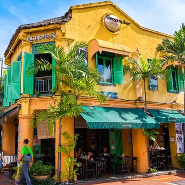 Yellow two-storey shophouse with green shutters, awnings and palms, and people at outdoor tables, in Melaka.
