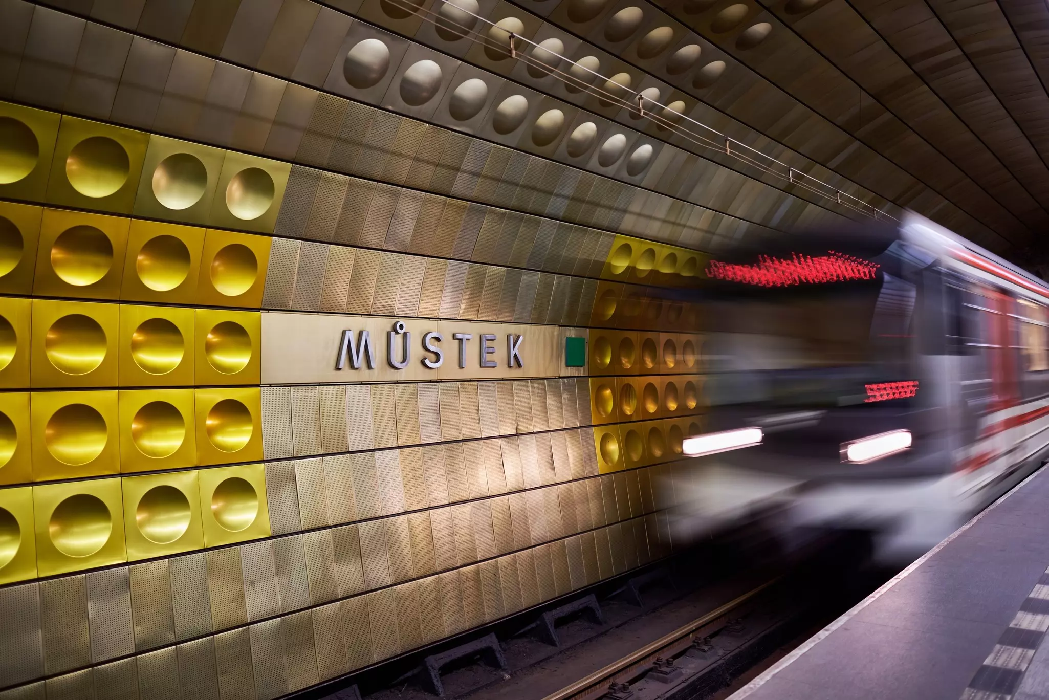 A subway train is blurry as it arrives in a metro station with a wall covered in aluminum panels