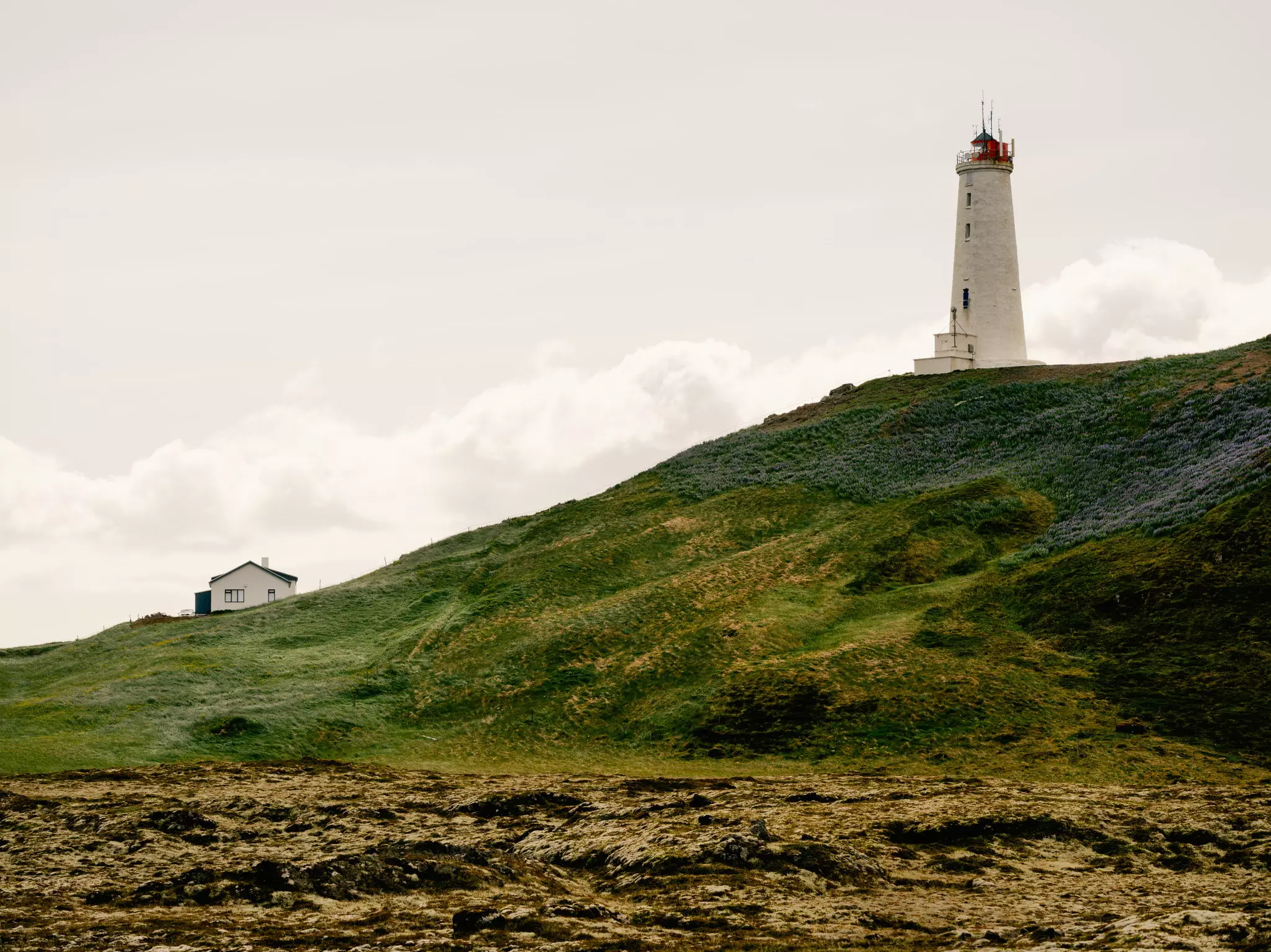 Lighthouse atop a grassy hillside with a white home at the bottom of the hill on a cloudy day.