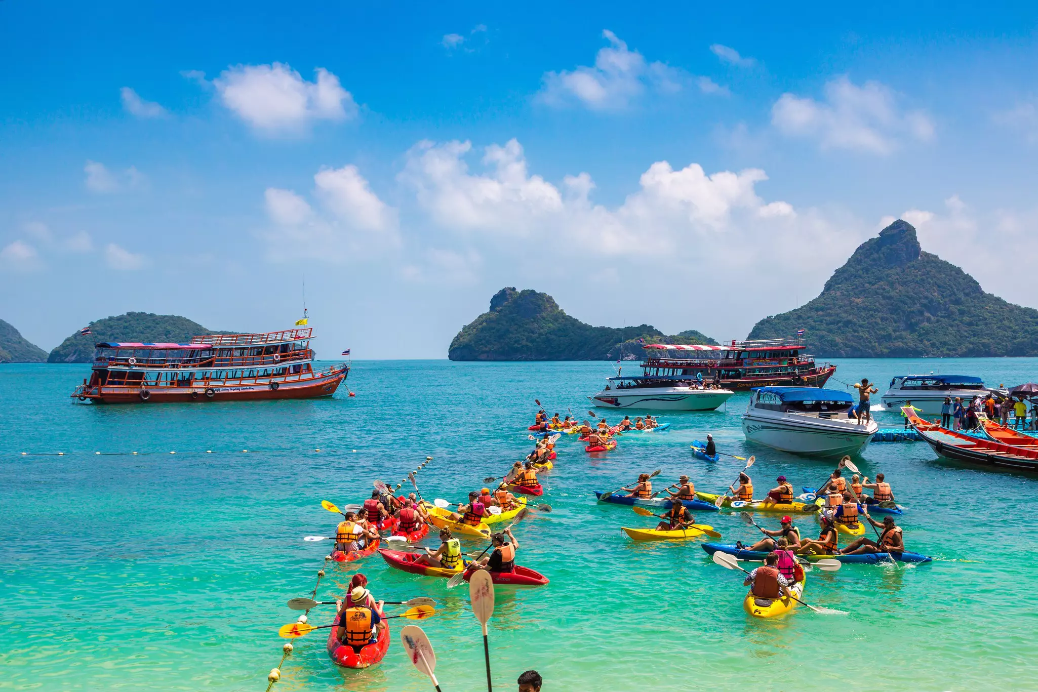 A dozen or so red and yellow kayaks are pictured in the turquoise waters off a tropical island. Boats and small, hilly islands are visible in the distance.