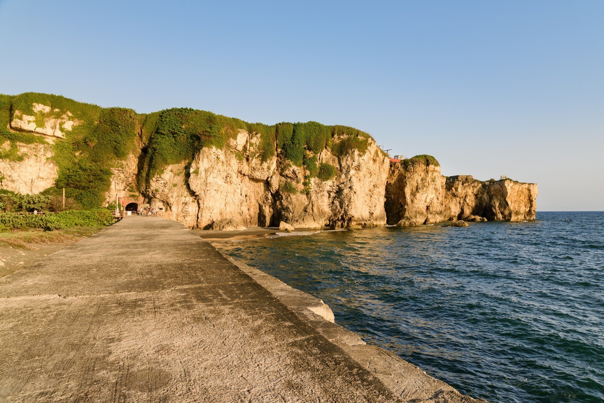 A concrete seawall path runs beside vine-draped limestone cliffs with a small tunnel opening and calm blue sea