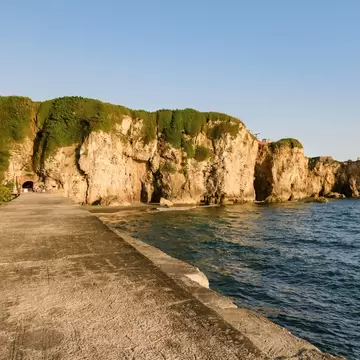 A concrete seawall path runs beside vine-draped limestone cliffs with a small tunnel opening and calm blue sea
