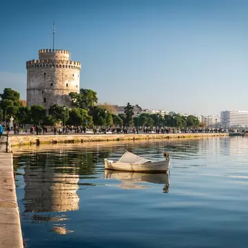 People stroll the seafront promenade beside the White Tower, with a rowing boat on calm water, in Thessaloniki, Greece