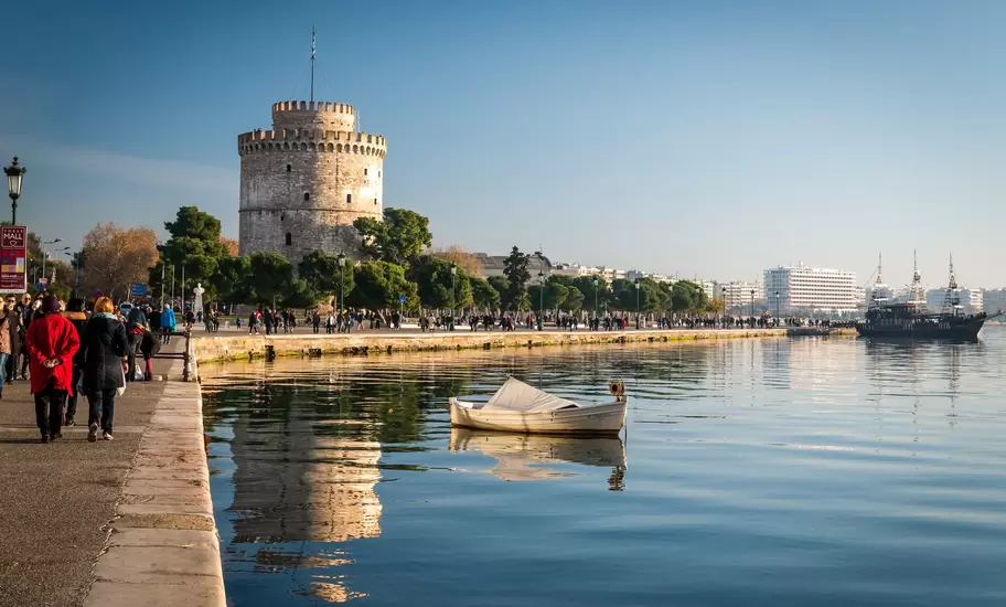 People stroll the seafront promenade beside the White Tower, with a rowing boat on calm water, in Thessaloniki, Greece