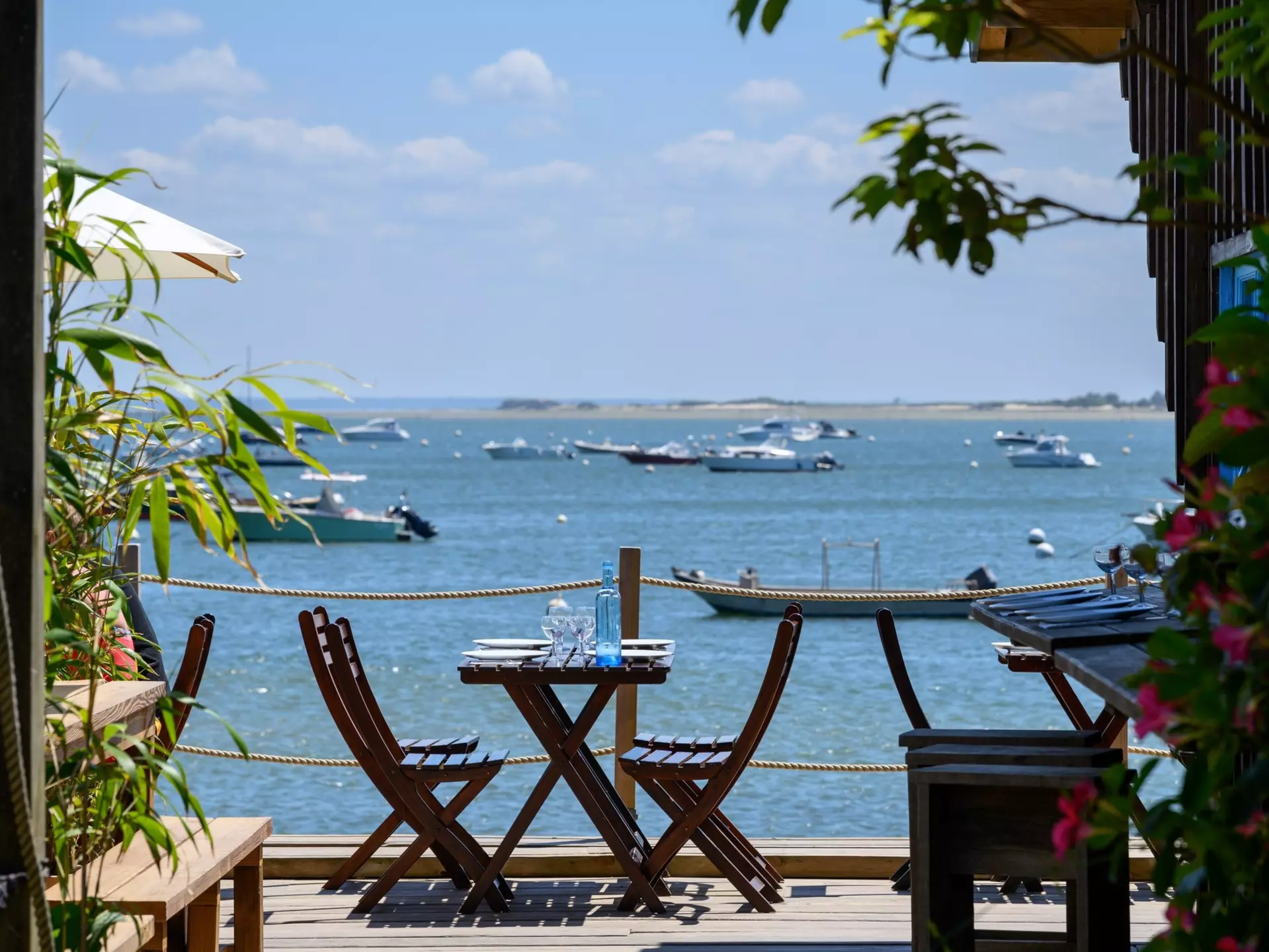 A table set on the decking of a casual seaside restaurant. Boats are docked in the bay.