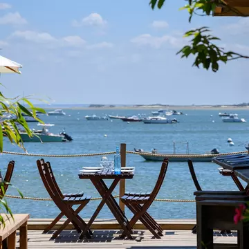 A table set on the decking of a casual seaside restaurant. Boats are docked in the bay.