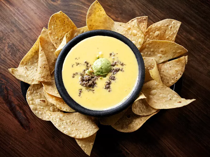 Bowl of yellow melted cheese with a scoop of guacamole, surrounded by corn chips.