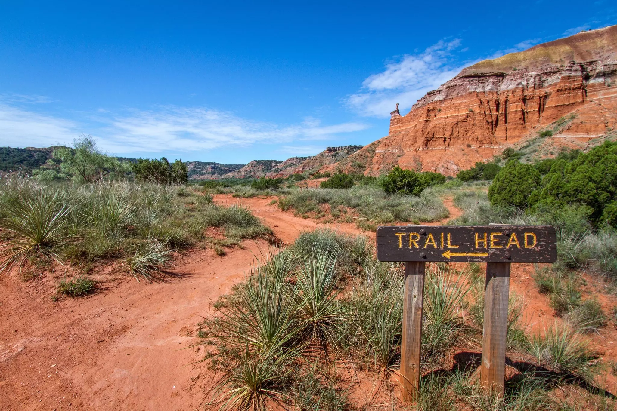 A sign leads toward a hiking trail in a desert landscape, with red, sandy soil, bushes and a rock formation in the distance.