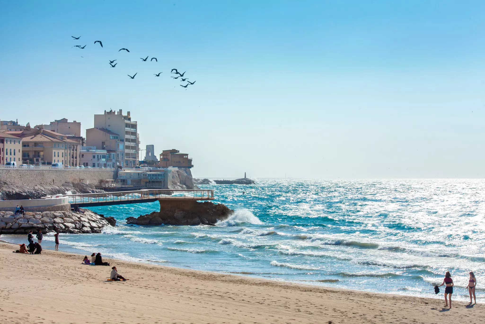 A sunny day on a sandy city beach. Waves crash on the shore and a few people sit on the sand enjoying the scene.