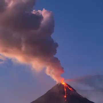 The cone of Mayon Volcano at dusk, a red lava stream at its peak and a large plume of ash rising into a blue sky.