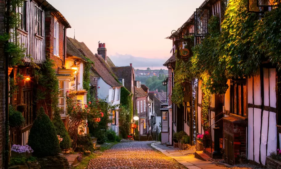 Cobbled Mermaid Street in Rye at dusk, lined with timber-framed and brick houses, ivy, roses and glowing lanterns