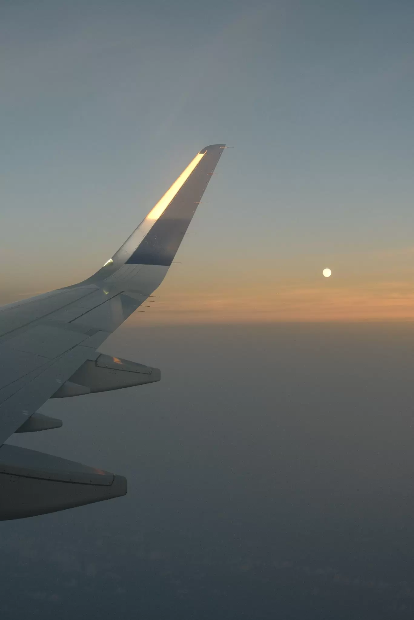 An airplane wing in flight and the moon at sunset.