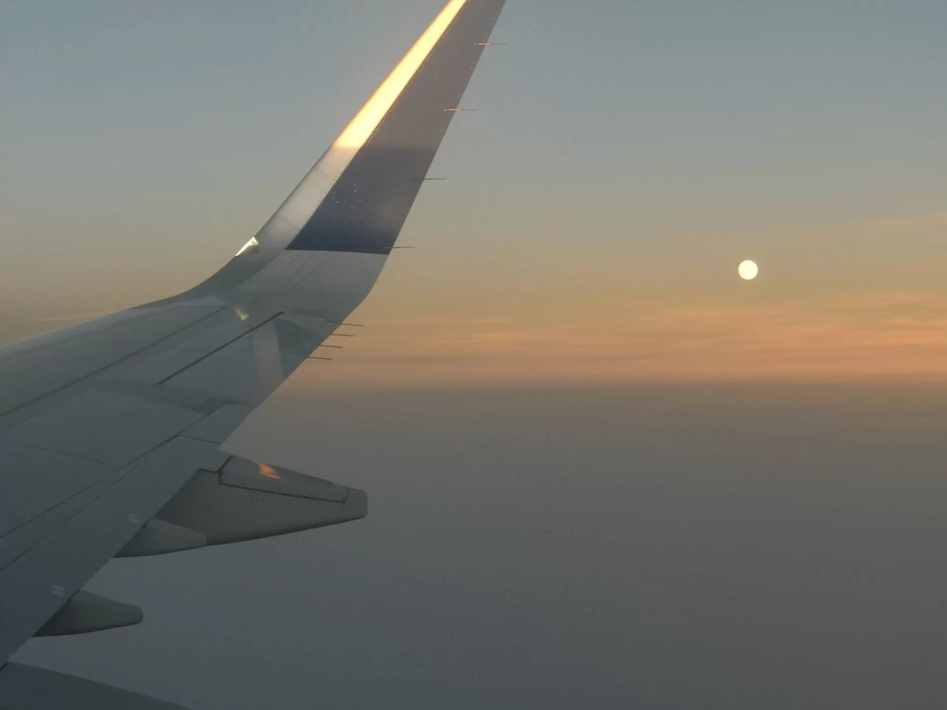 An airplane wing in flight and the moon at sunset.