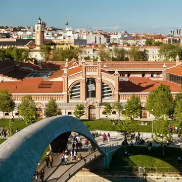 Locals and tourists walk though a modern bridge over the Manzanares River in the public park of Madrid Río in Madrid, Spain, with the Matadero building (a former slaughterhouse converted to an arts centre) in the background.