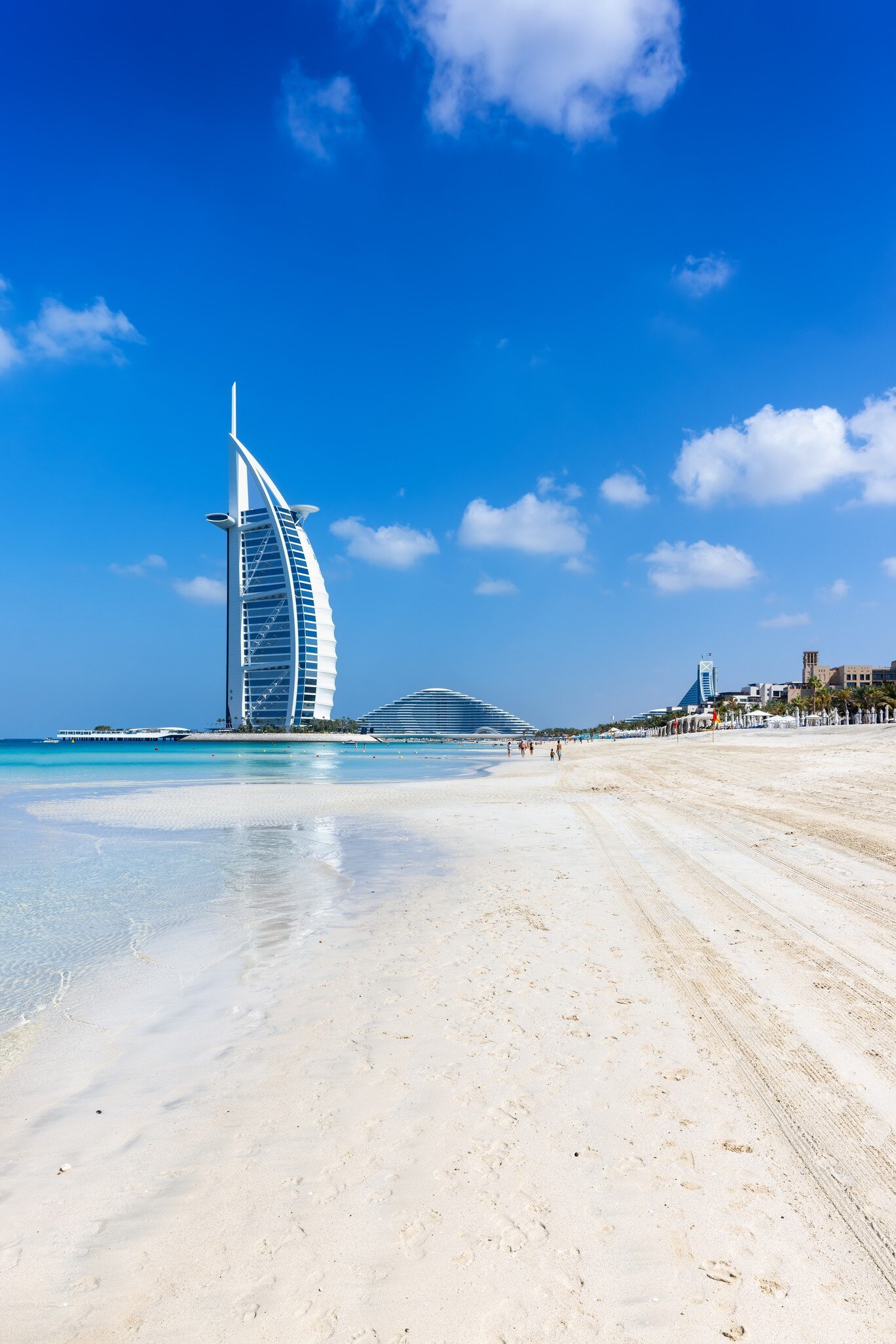 A sail-shaped hotel standing offshore beyond a wide empty beach.