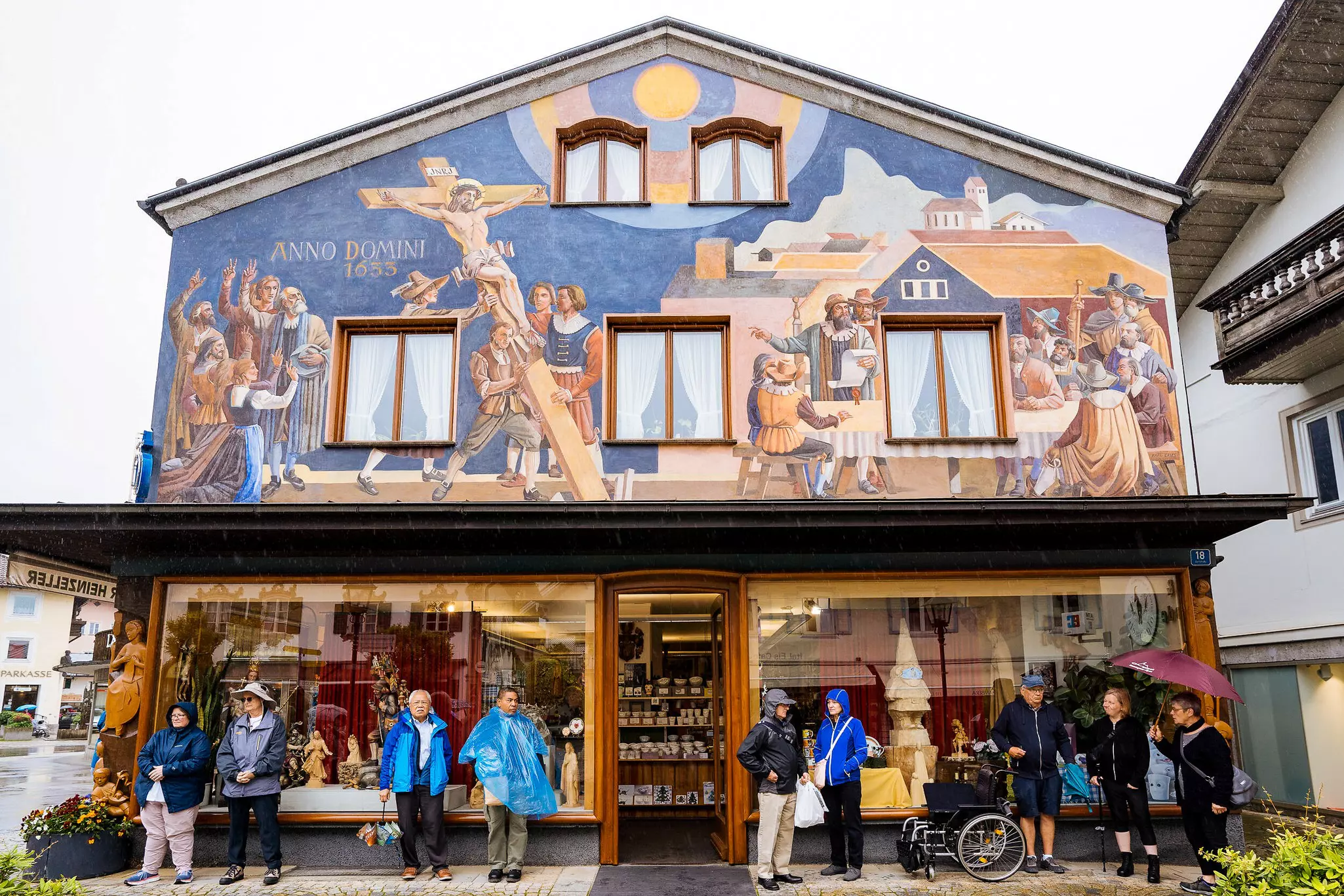 Tourists hide from the rain under the awning of a building with a painted fresco of the village's historic Passion Play