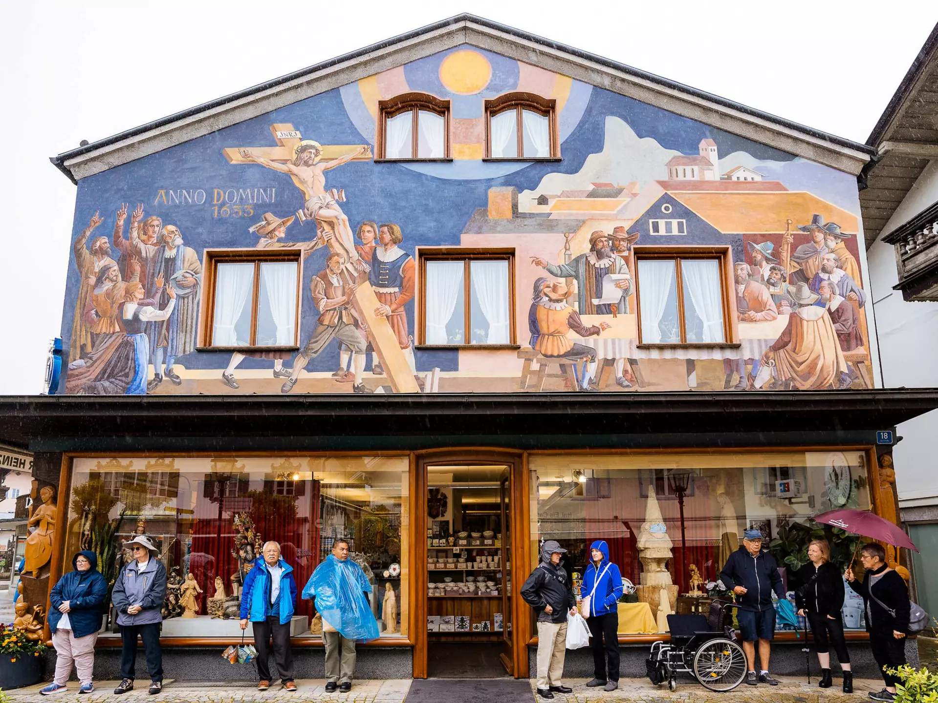 Tourists hide from the rain under the awning of a building with a painted fresco of the village's historic Passion Play