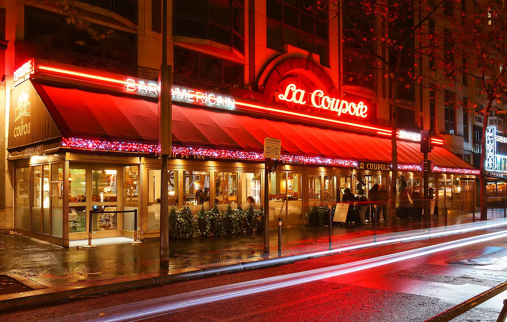 A brasserie in Paris, its exterior illuminated by neon, is seen from the street at night.