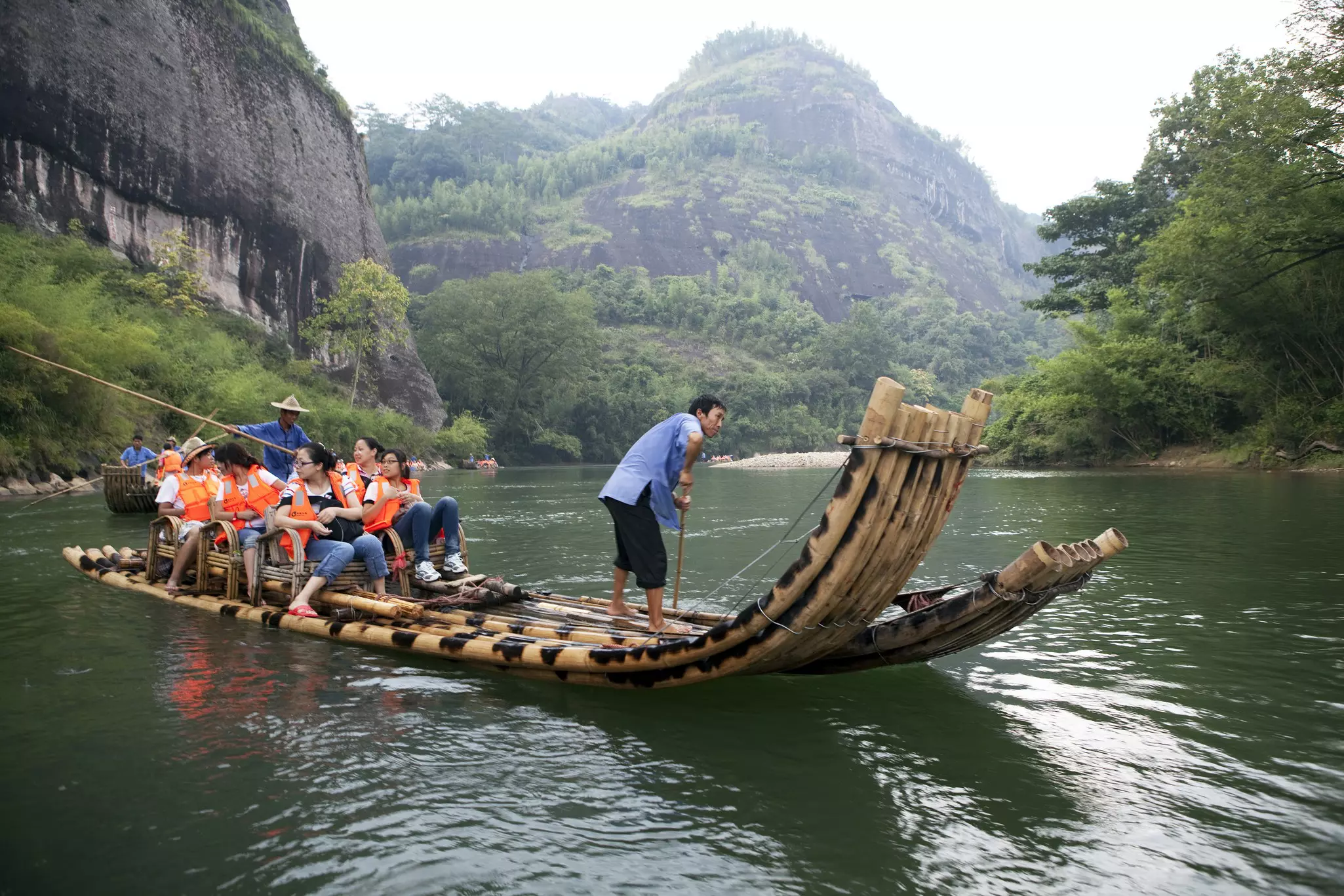 A bamboo boat pilot pushes a tourist boat along a river, past steep mountains.