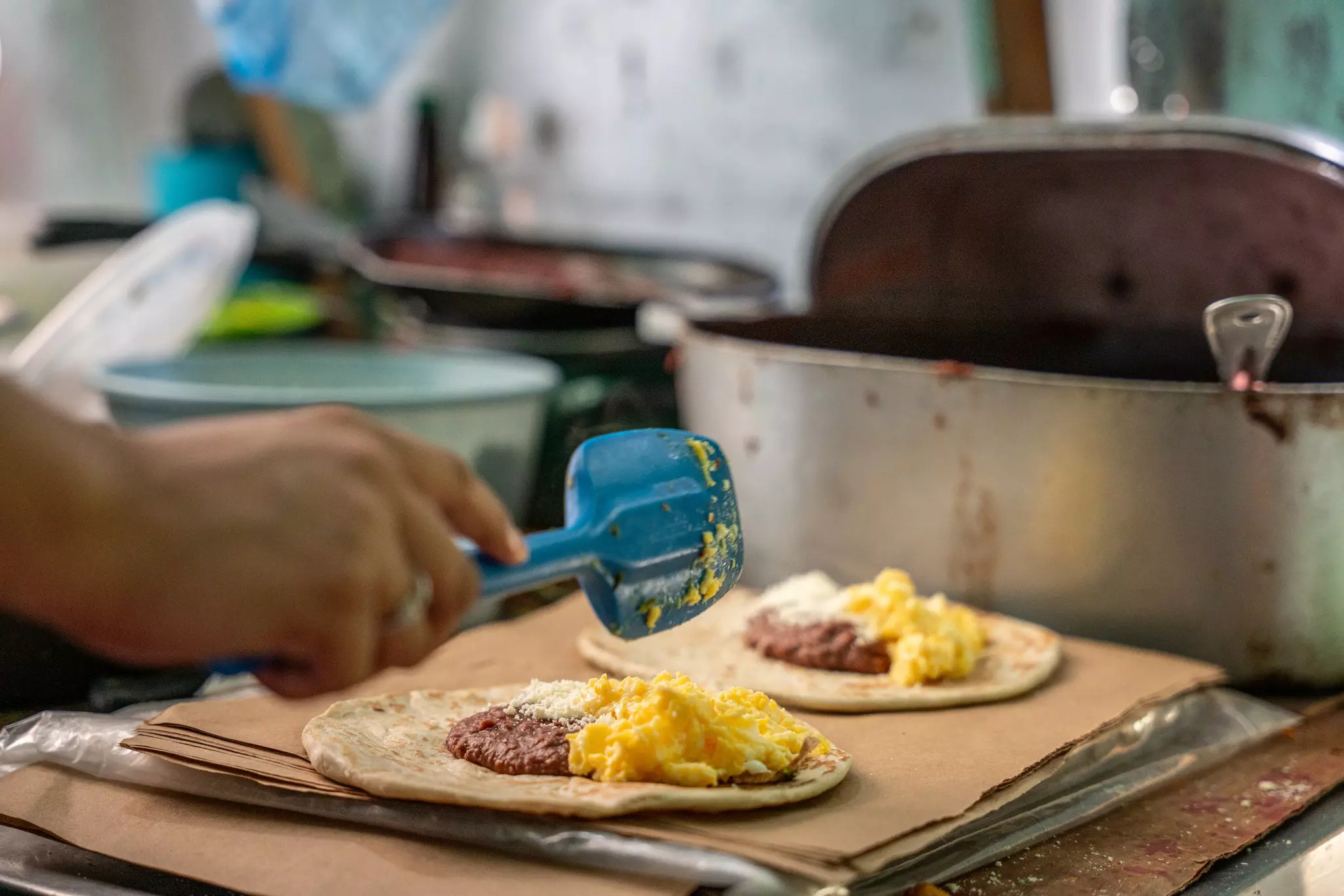 A person's hand fills tortillas with beans and eggs on a commercial kitchen workspace.