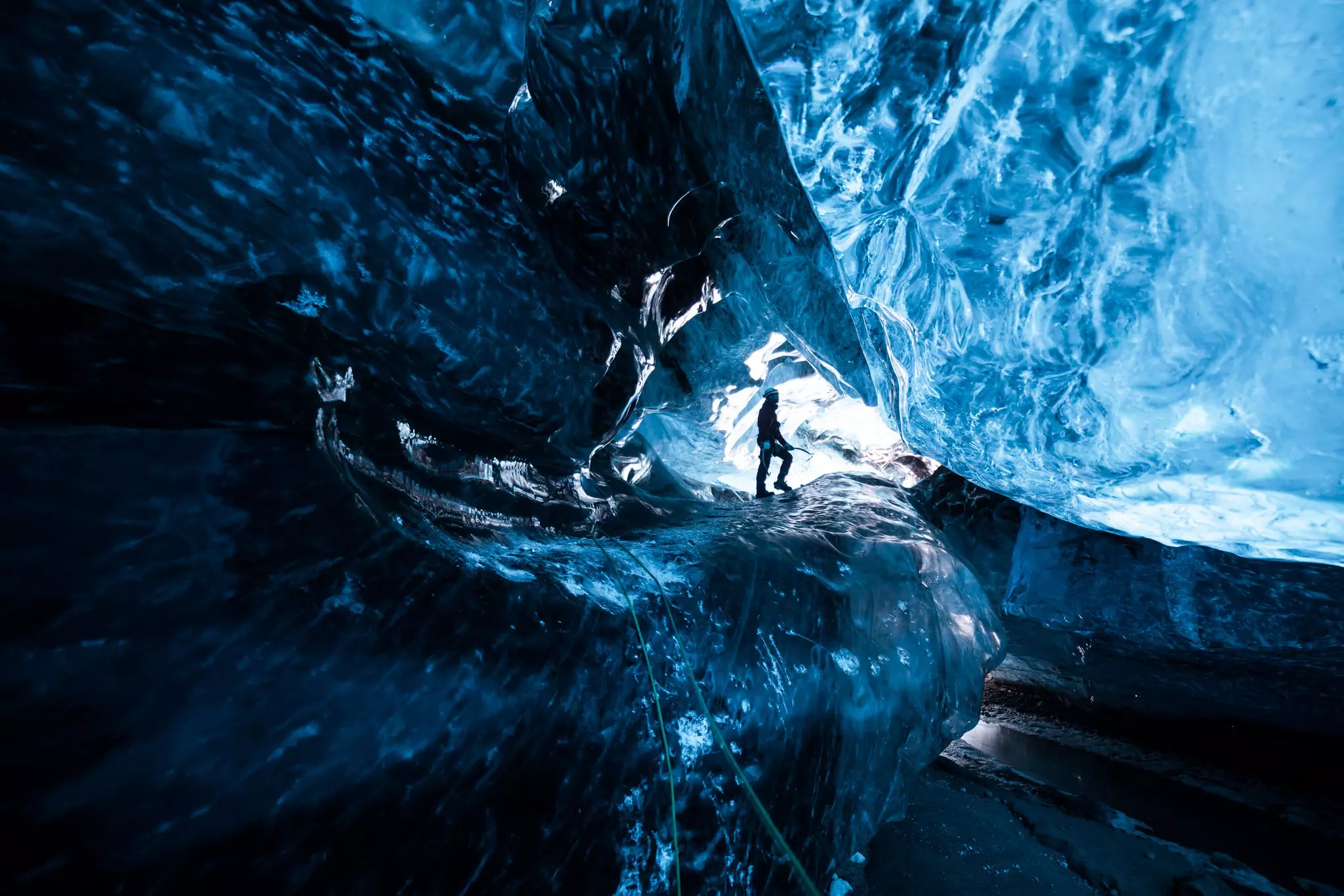 A person is silhouetted inside an ice cave. The sculptural sides of the cave seem to glow blue in the light.